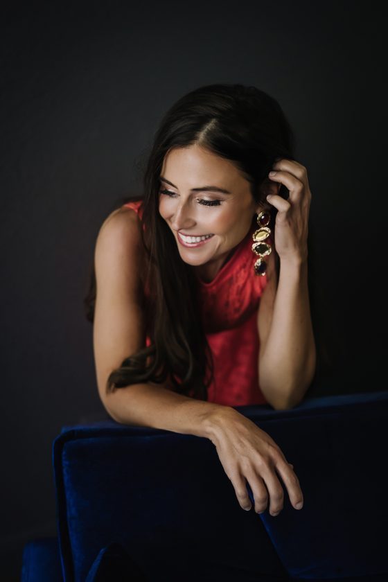 Solara Rose in a coral dress and gemstone drop earrings, eyes closed and smiling, leaning on a blue velvet chair